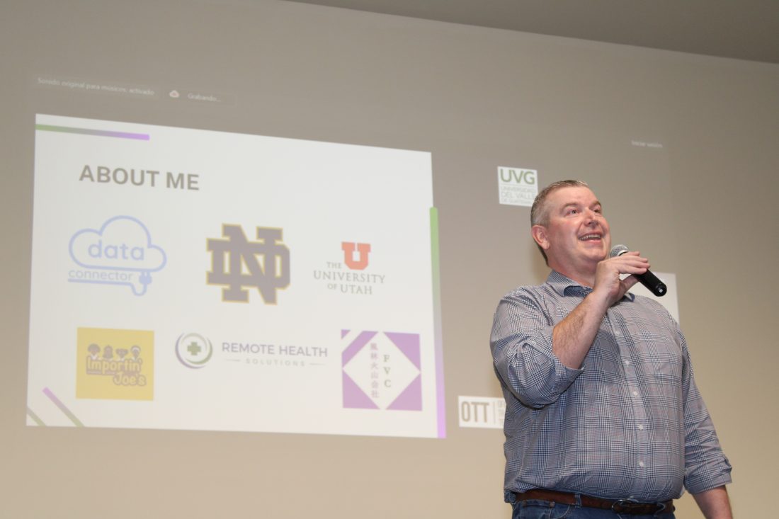 James Thompson speaking in front of group on stage with a PowerPoint projected behind him