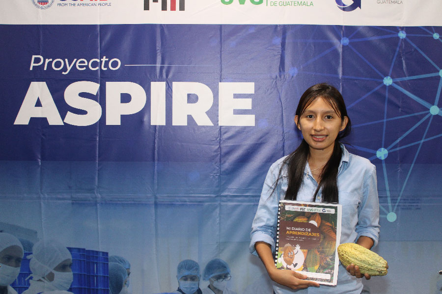 Mujer mostrando Diario de Aprendizaje frente a banner del proyecto ASPIRE. 