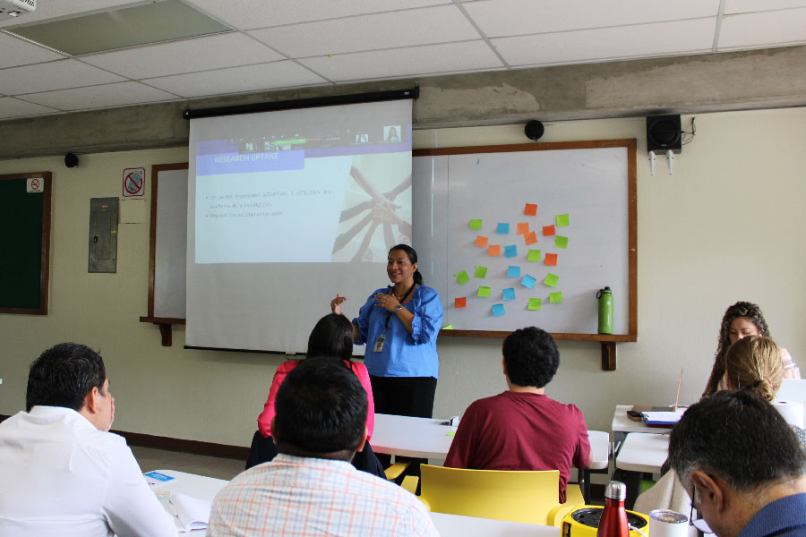 Mujer hablando frente a un grupo de personas en un aula. 